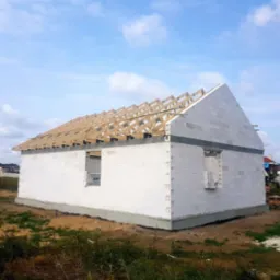 Surowy stan domu jednorodzinnego z białych bloczków, widoczna drewniana konstrukcja dachu bez pokrycia, okna jeszcze niezamontowane.