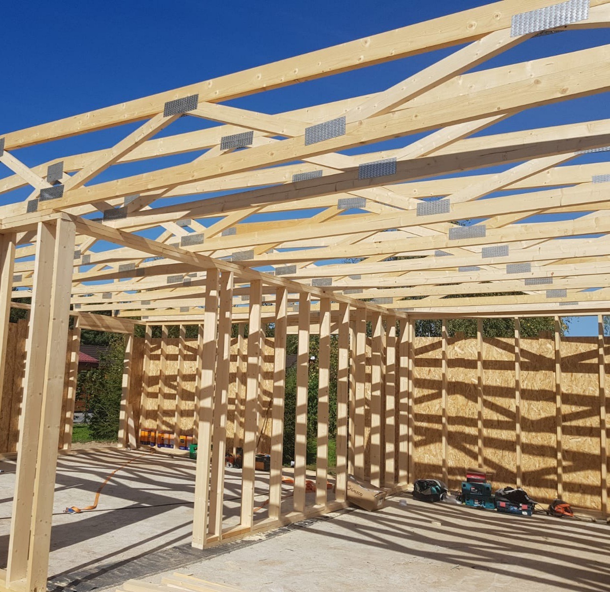 Surowa konstrukcja drewniana dachu z widocznymi elementami łączeniowymi z metalu, w trakcie budowy, z narzędziami i materiałami budowlanymi leżącymi na podłodze.