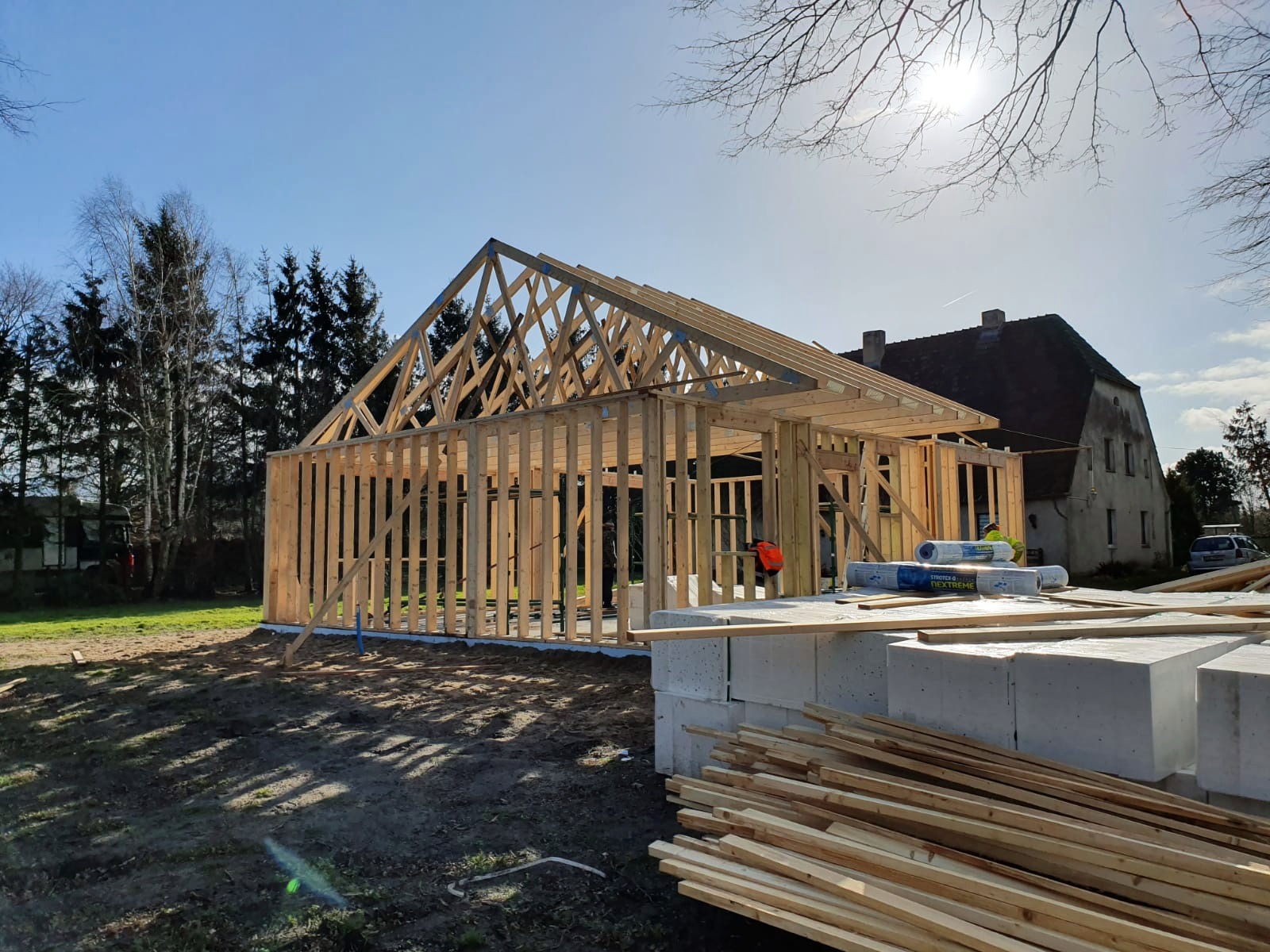 Surowa konstrukcja drewniana domu w trakcie budowy, widoczne elementy szkieletu, bloczki betonowe i rolki izolacji, w tle budynek mieszkalny i drzewa.