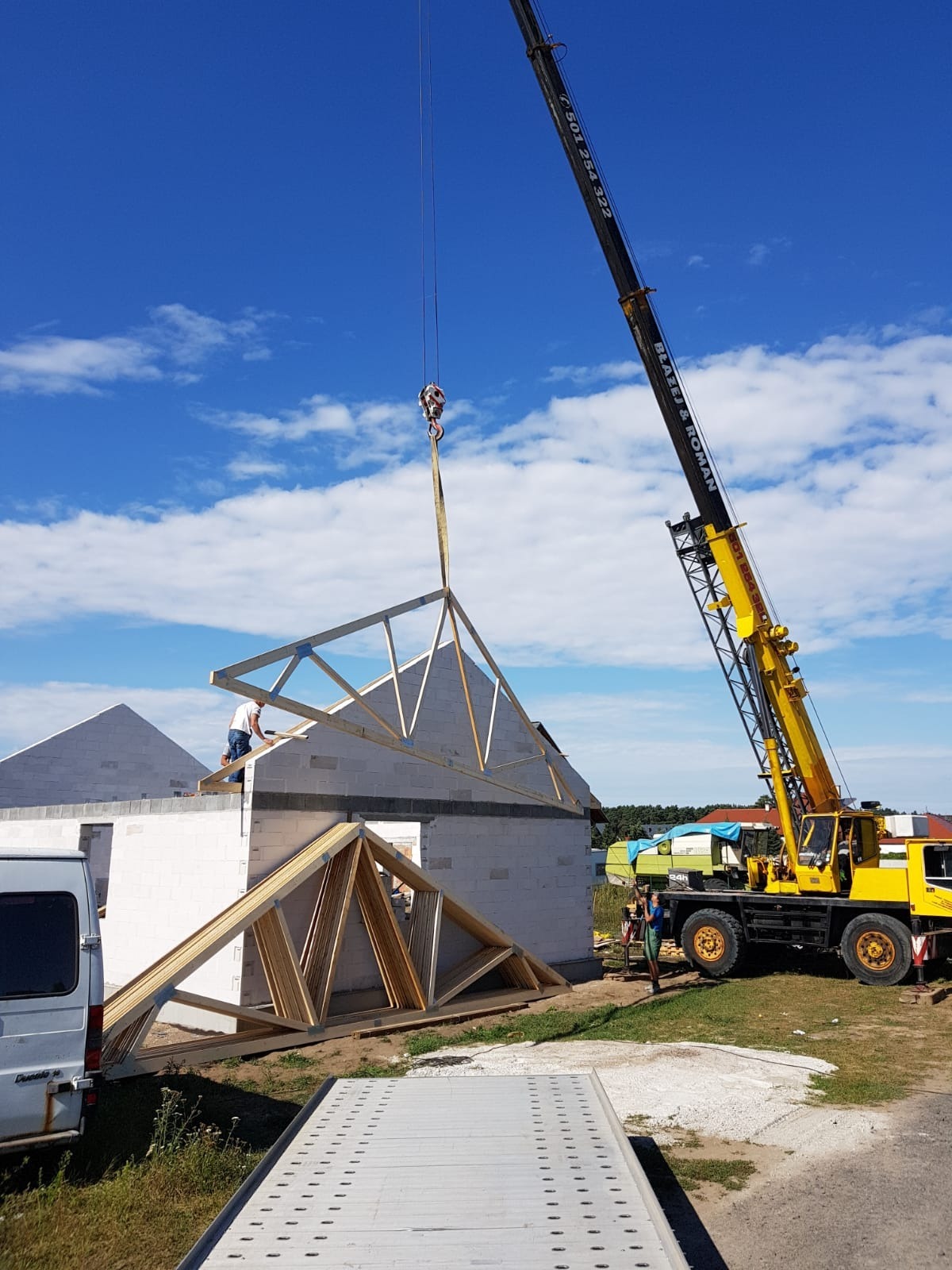 Żółty dźwig unosi drewnianą konstrukcję dachu nad budynkiem z białych bloczków, pracownik na ścianie, błękitne niebo w tle.
