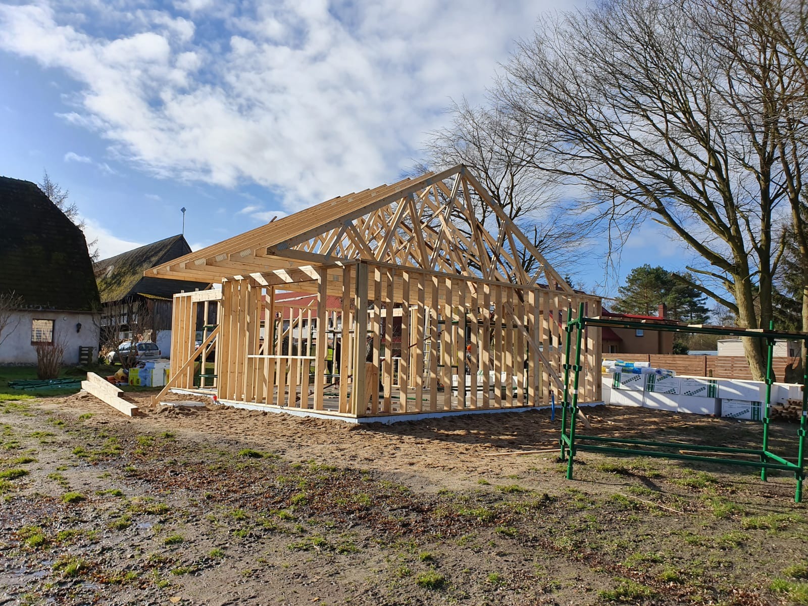 Wznoszenie drewnianej konstrukcji szkieletowej budynku z widoczną więźbą dachową w trakcie montażu, na tle błękitnego nieba z chmurami i drzew bez liści.