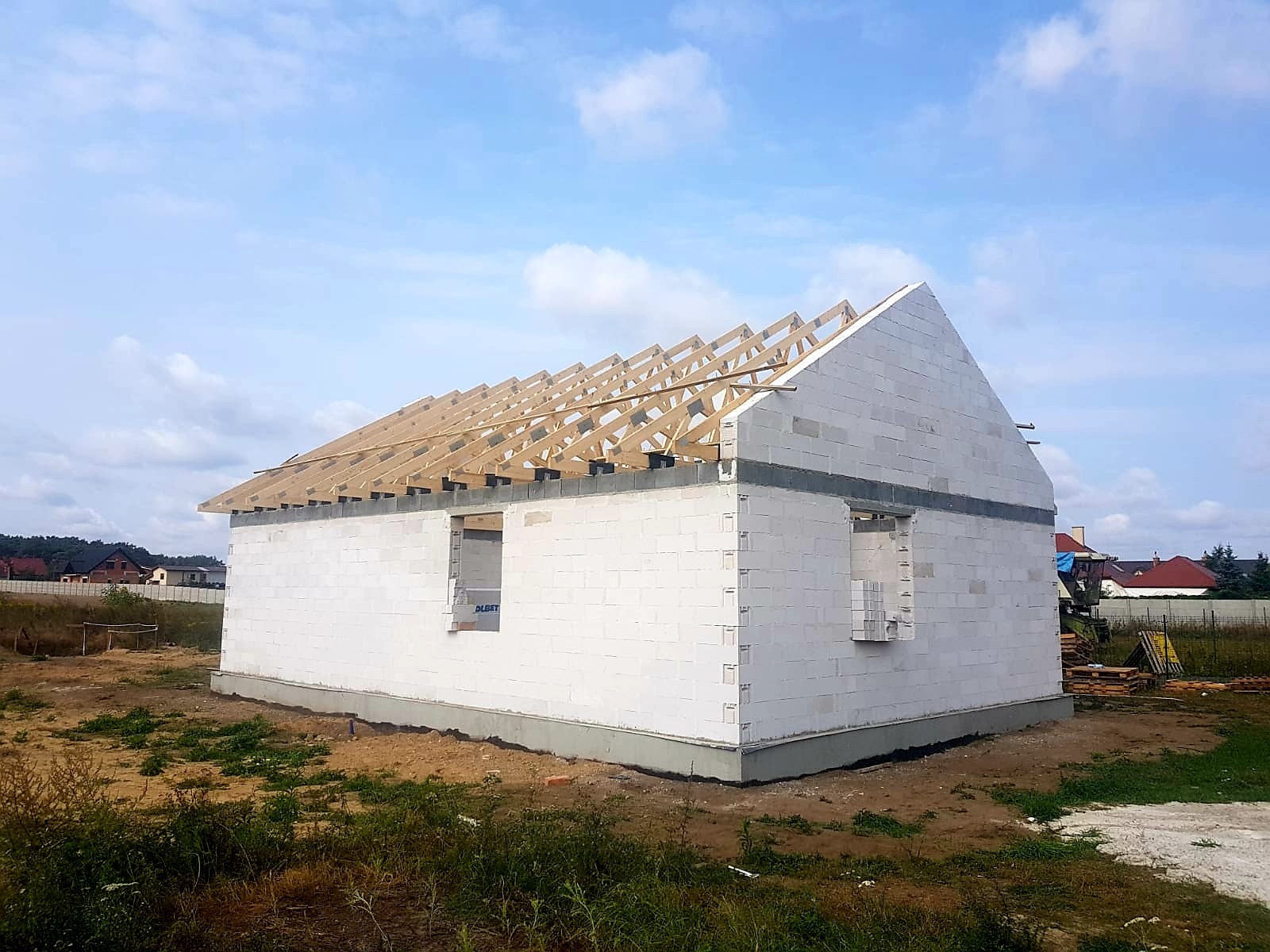 Surowy stan domu jednorodzinnego z białych bloczków, widoczna drewniana konstrukcja dachu bez pokrycia, okna jeszcze niezamontowane.