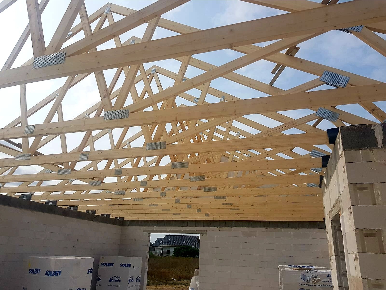 Konstrukcja drewniana dachu w trakcie montażu, widoczne metalowe łączniki, ściany z białych bloczków, niebo w tle.