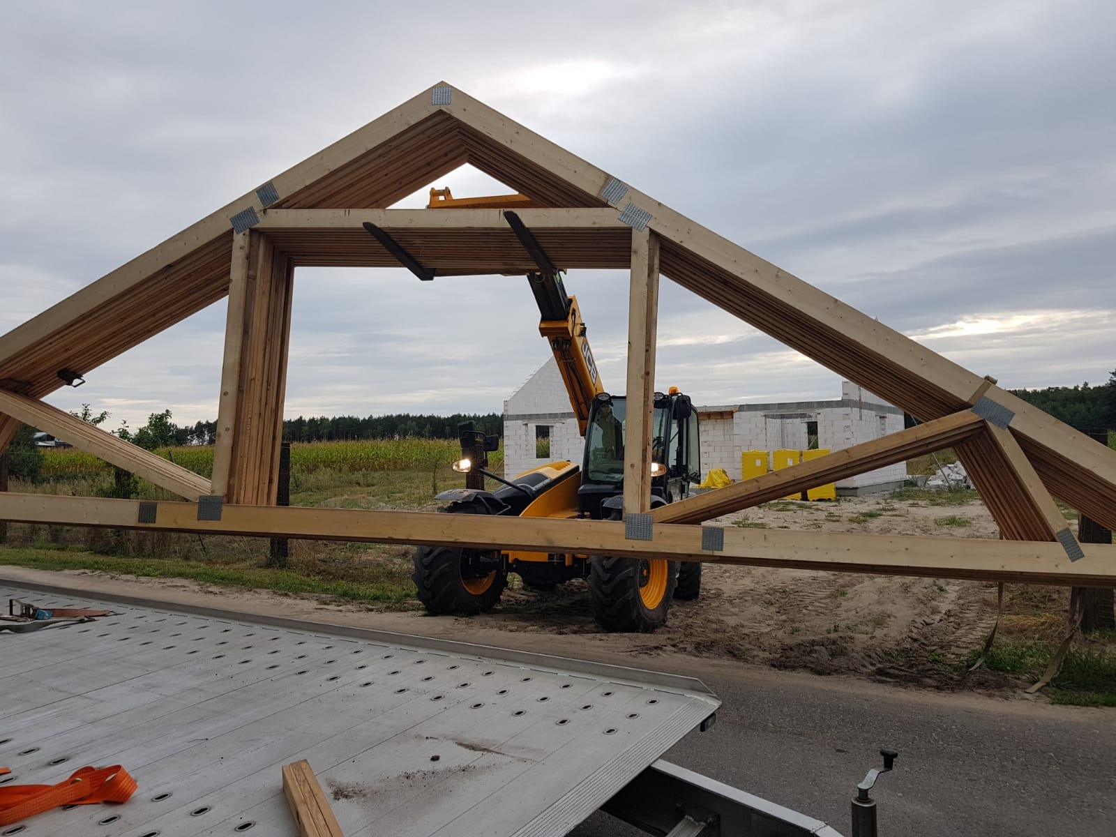 Żuraw teleskopowy podnosi gotowy element drewnianej konstrukcji dachowej, widoczny budynek w trakcie budowy.
