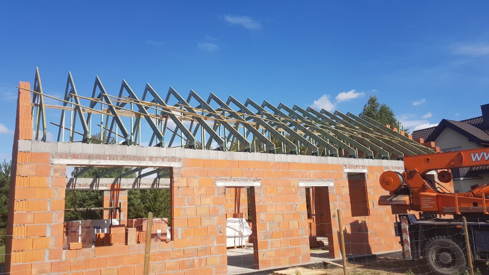 Wznoszona więźba dachowa na budynku z czerwonej cegły, widoczny dźwig budowlany w tle, błękitne niebo.