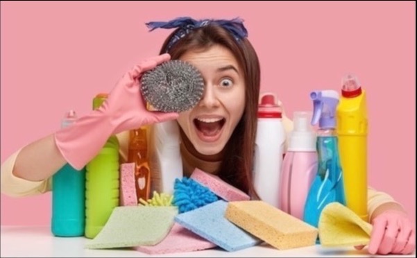 Uśmiechnięta kobieta w różowych rękawicach trzyma drucianą myjkę przed okiem, otoczona butelkami środków czystości i gąbkami na różowym tle.