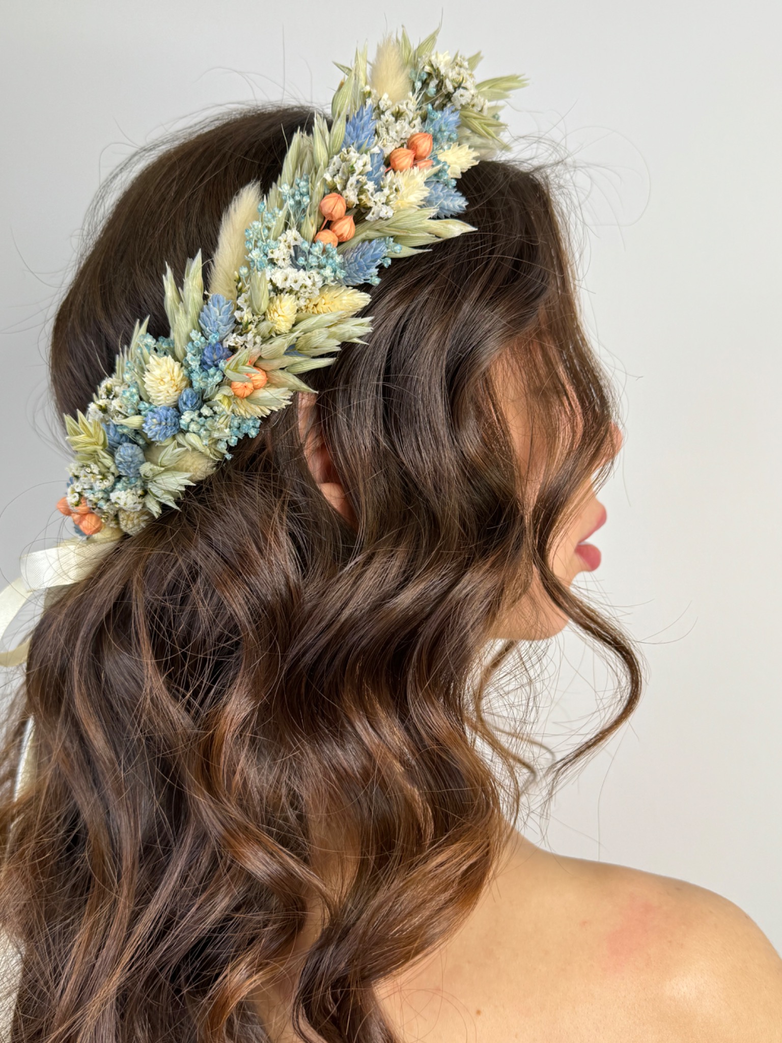Profil kobiety z długimi, brązowymi, kręconymi włosami, noszącej delikatny wianek z suszonych kwiatów w odcieniach błękitu, kremu i pomarańczy, z białymi wstążkami.