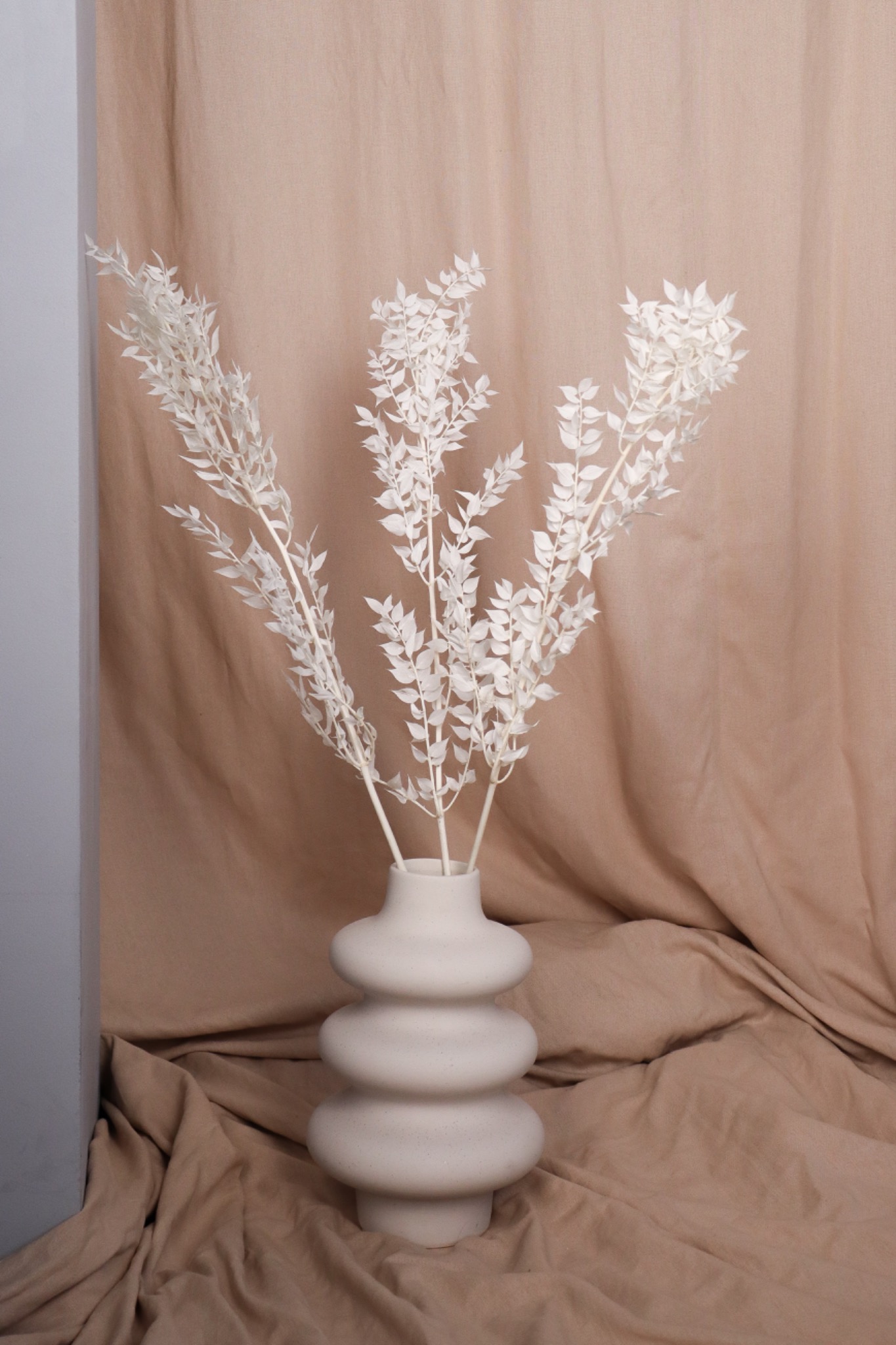 Beżowa, ceramiczna waza o obłym kształcie z trzema białymi gałązkami na tle drapowanej tkaniny w odcieniach beżu.