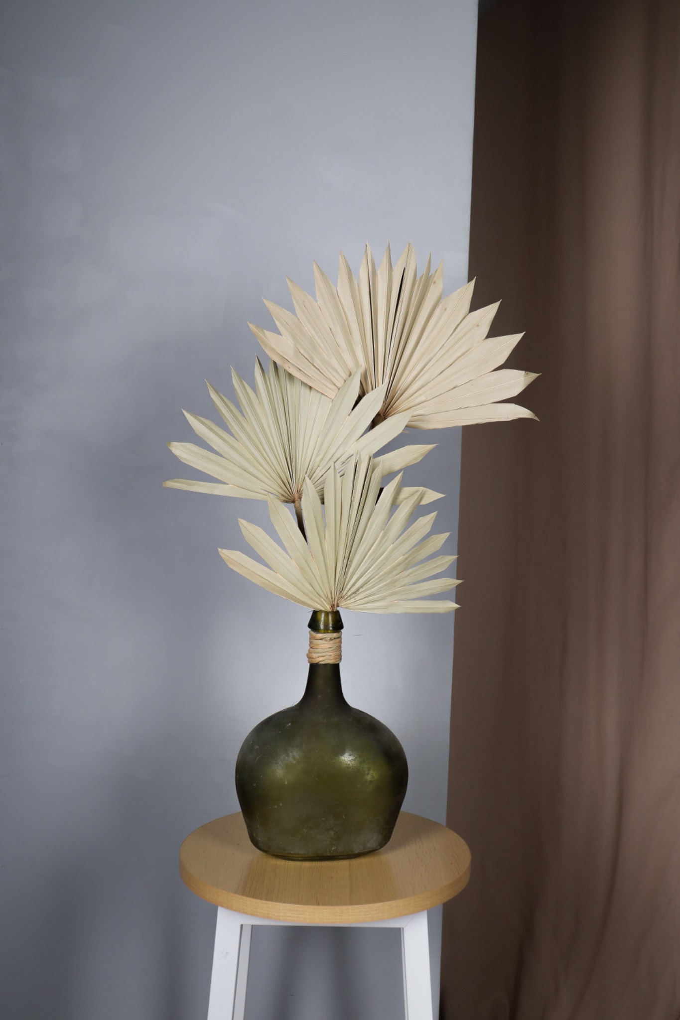 Kompozycja z suszonych liści palmowych w zielonej, obwiązanej sznurkiem butelce na okrągłym, drewnianym stołku z białymi nogami, na tle szarej i brązowej tkaniny.
