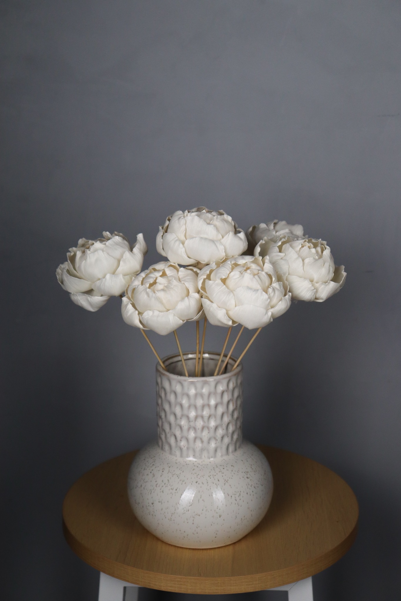 Kompozycja sześciu białych, ręcznie robionych kwiatów umieszczonych w ceramicznym wazonie o teksturowanej powierzchni, stojącym na okrągłym, drewnianym stoliku na tle szarej ściany.