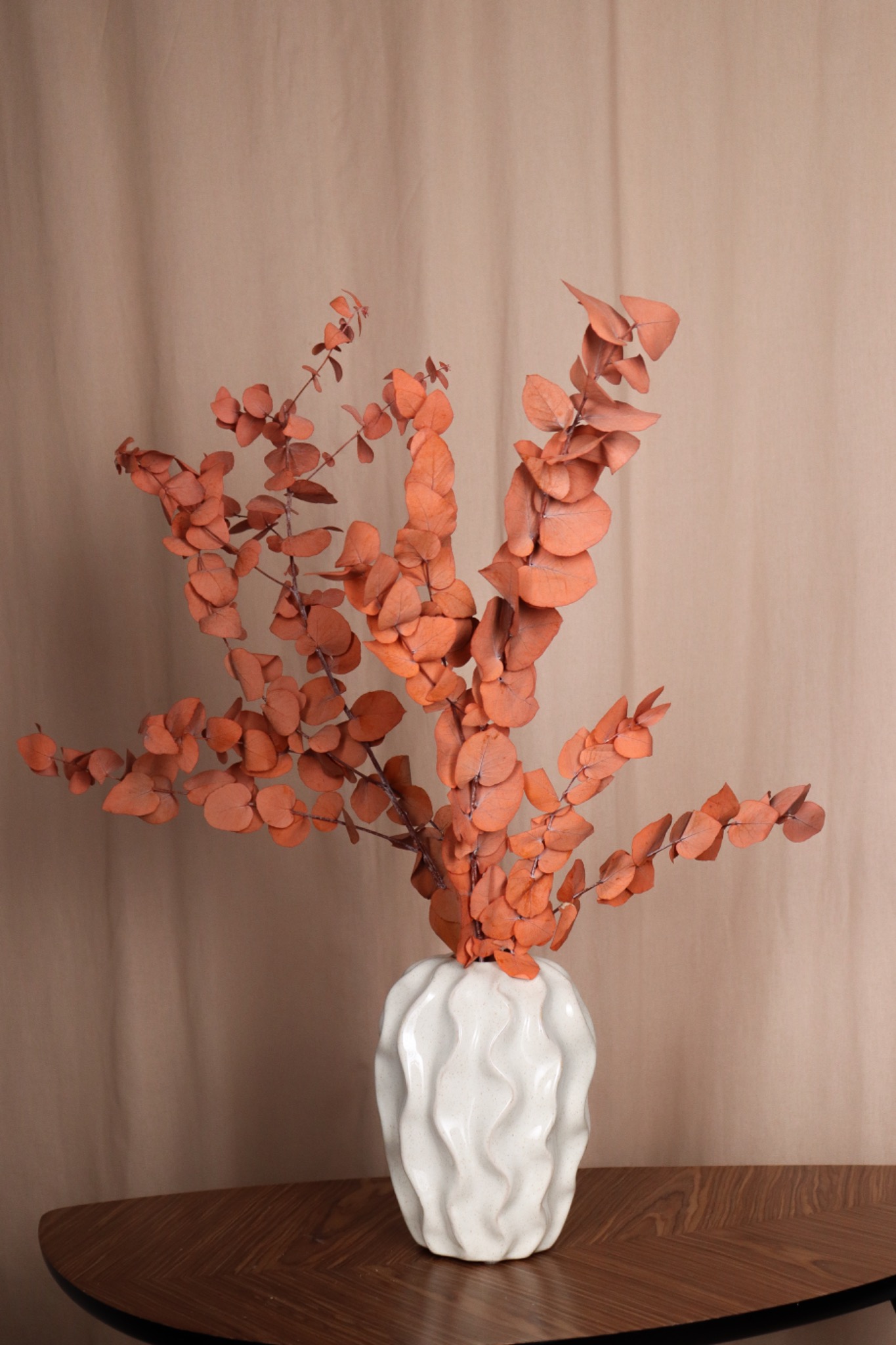 Kompozycja z suszonego eukaliptusa o miedzianych liściach w białym, ceramicznym wazonie z falistą fakturą, ustawiona na drewnianym stole o wzorzystym blacie na tle beżowej tkaniny.