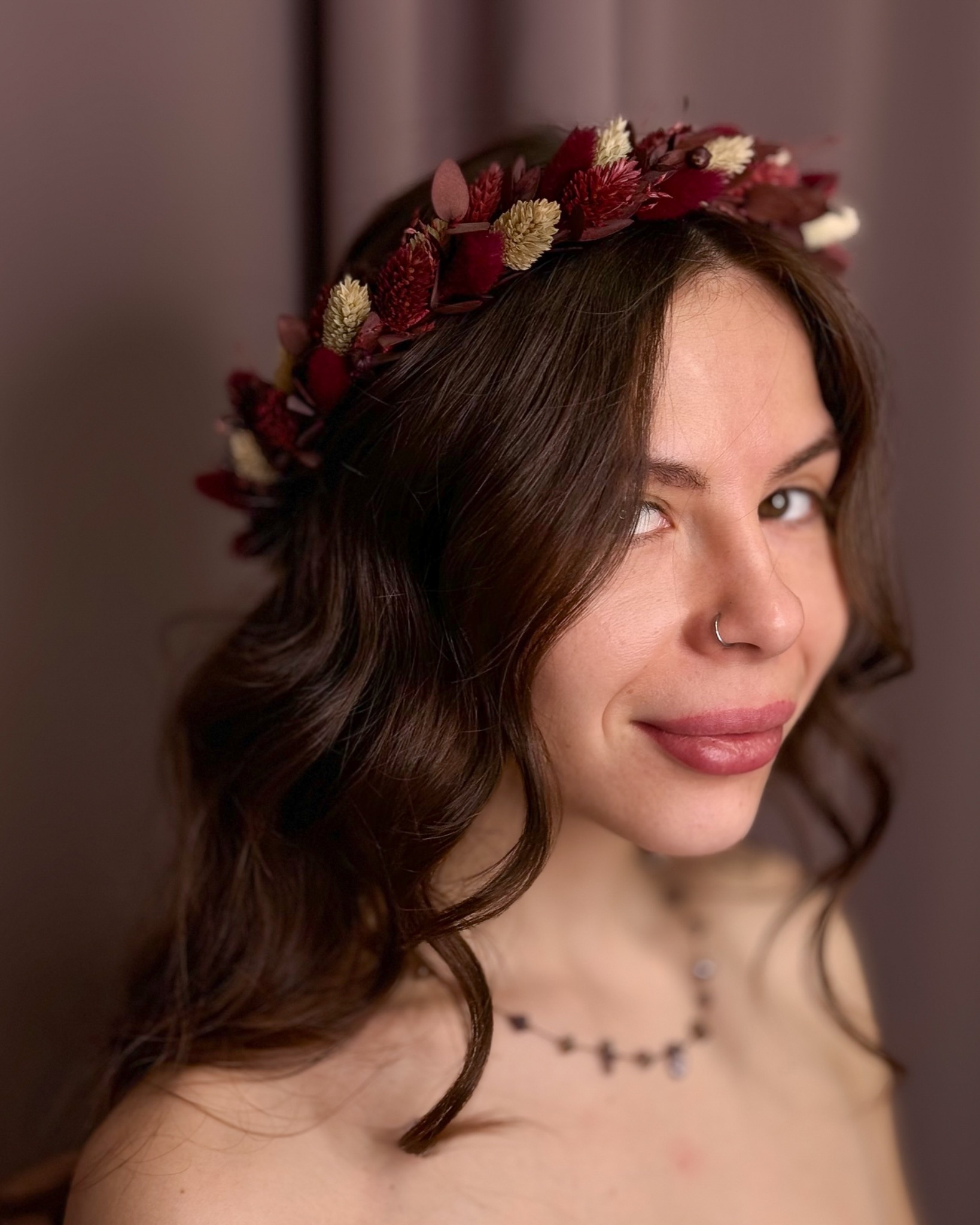 Portret kobiety z ciemnymi, kręconymi włosami i wiankiem z suszonych kwiatów w odcieniach bordo i beżu na głowie, z delikatnym makijażem i kolczykiem w nosie, uśmiechająca się do kamery.