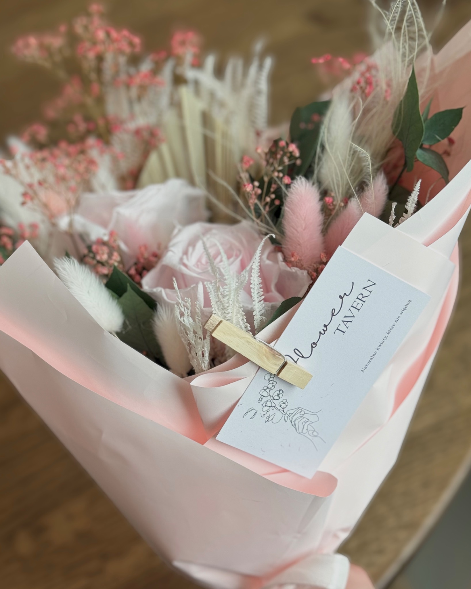 Delikatny bukiet w pastelowych odcieniach różu i bieli, z dodatkiem suszonych kwiatów i piórek, zapakowany w różowy papier, z dołączoną wizytówką Flower Tavern, spiętą drewnianym klamerką.
