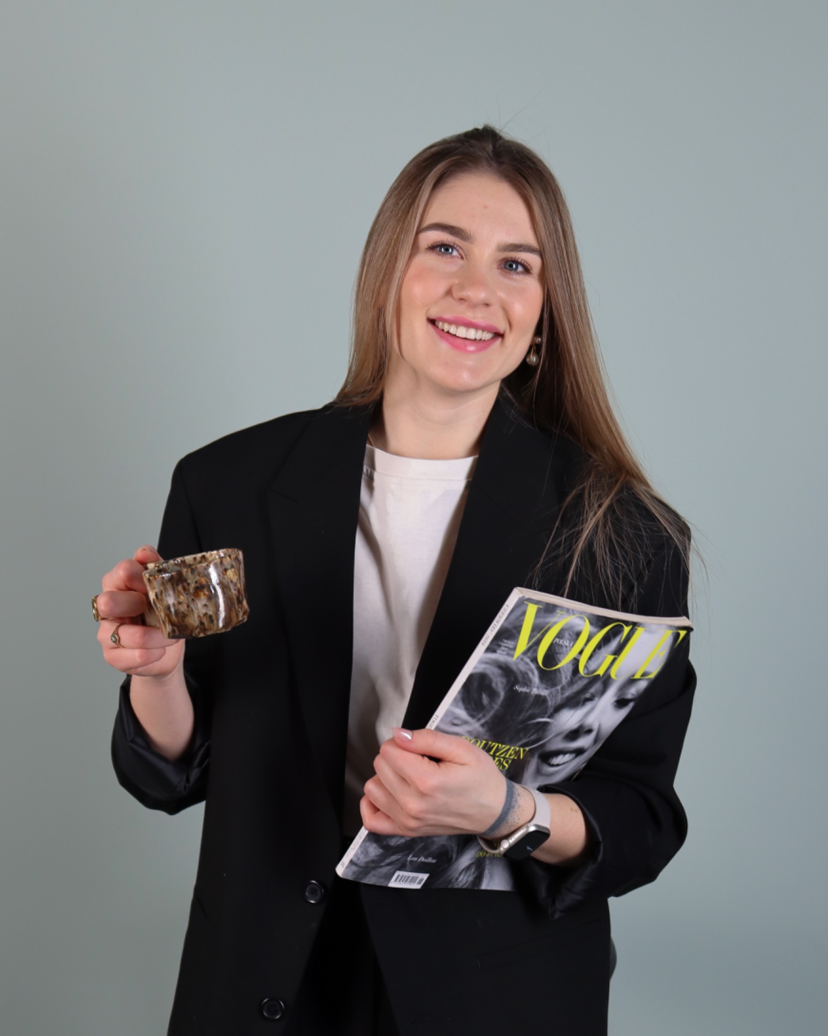 Uśmiechnięta kobieta w czarnej marynarce trzyma kubek z wzorem i magazyn Vogue, na palcu złoty pierścionek.