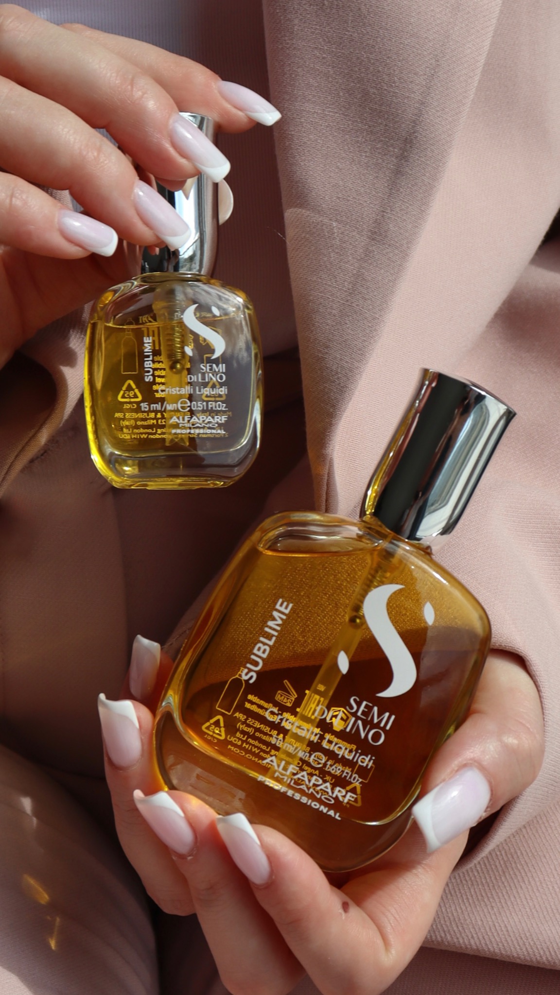 Dwie buteleczki olejku do włosów Semi Di Lino w dłoniach kobiety z french manicure na tle pudrowego różu marynarki.