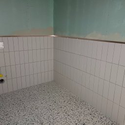 Ihor Petrachenko - Ściany w łazience wyłożone białymi, wąskimi płytkami, ułożonymi pionowo, z metalową listwą na górze, oraz podłoga z lastryko w odcieniach szarości. Widoczny otwór kanalizacyjny z żółtą zaślepką.