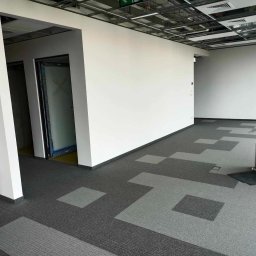 Ihor Petrachenko - Wnętrze biurowe po świeżym malowaniu ścian na biało, z szaro-grafitową wykładziną w geometryczny wzór i widoczną instalacją sufitową.