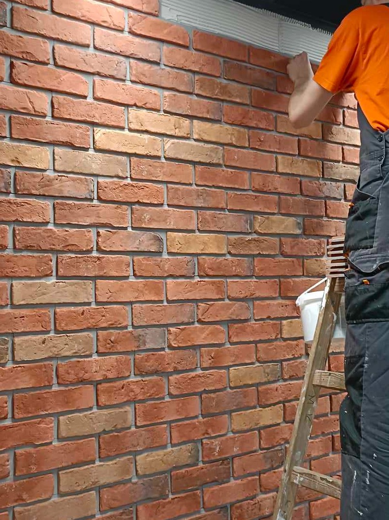 Montaż dekoracyjnej ściany z cegły, widoczna drabina, wiadro z klejem i fragment osoby w pomarańczowej koszulce i czarnym kombinezonie.