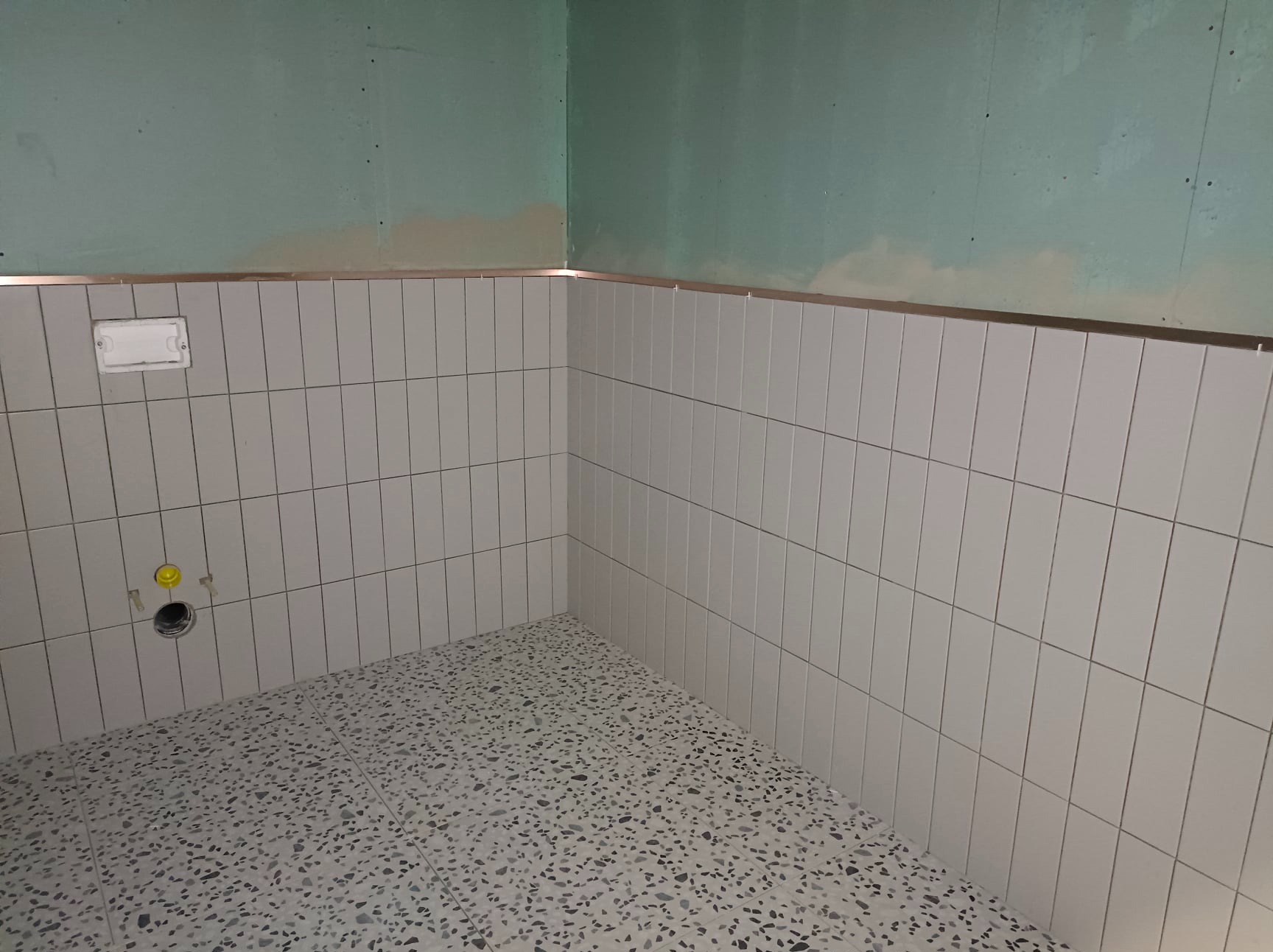 Ściany w łazience wyłożone białymi, wąskimi płytkami, ułożonymi pionowo, z metalową listwą na górze, oraz podłoga z lastryko w odcieniach szarości. Widoczny otwór kanalizacyjny z żółtą zaślepką.