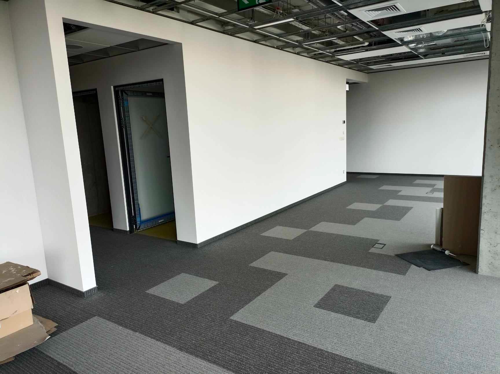 Wnętrze biurowe po świeżym malowaniu ścian na biało, z szaro-grafitową wykładziną w geometryczny wzór i widoczną instalacją sufitową.