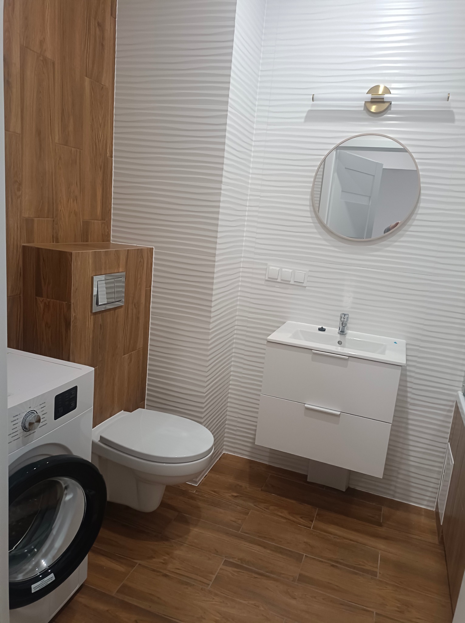 Nowoczesna łazienka z drewnopodobną podłogą i białymi, falistymi płytkami ściennymi. Umywalka z szafką, okrągłe lustro i pralka w rogu.