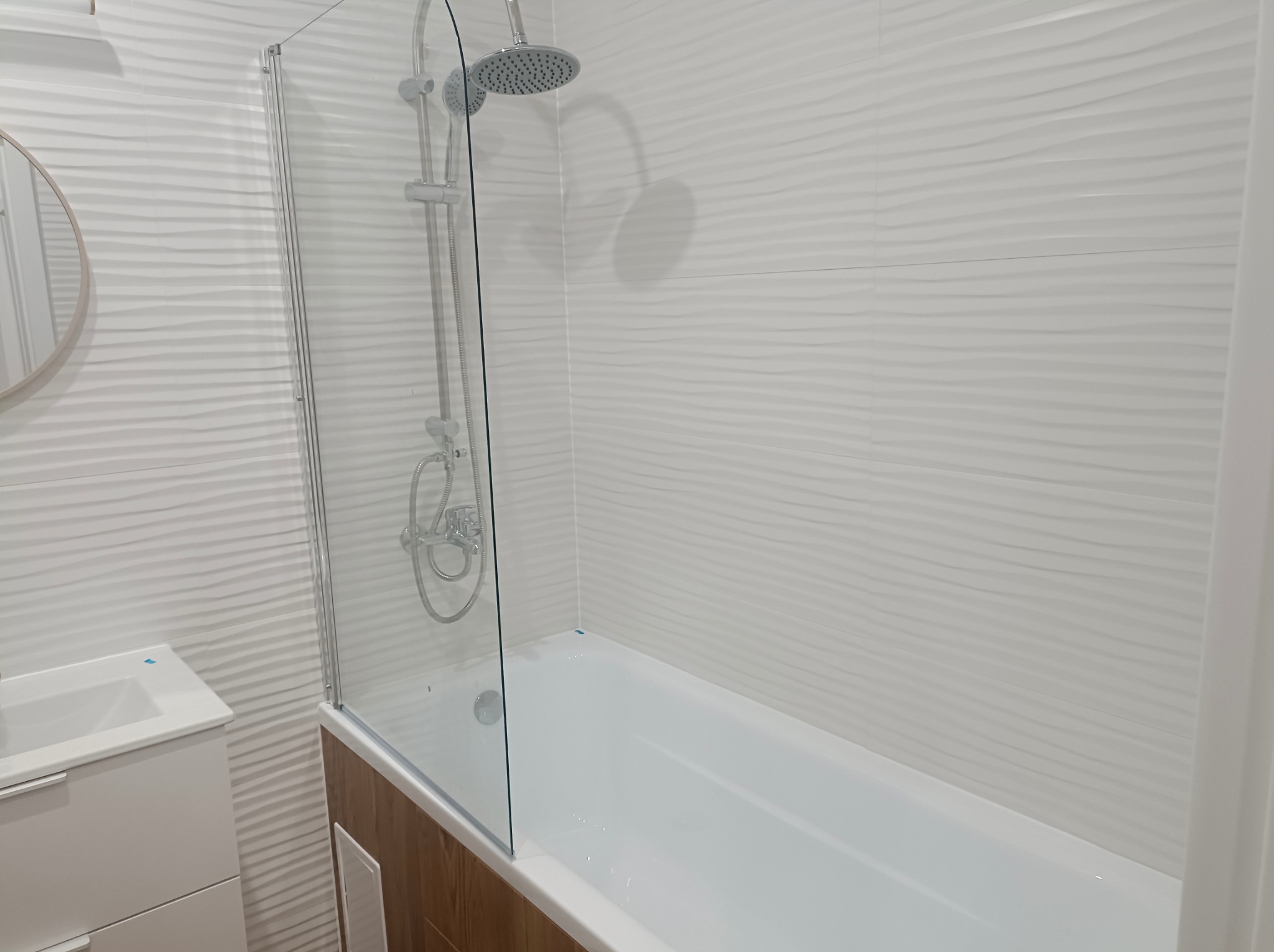 Nowoczesna łazienka z białą wanną, szklanym parawanem nawannowym i umywalką. Ściany wyłożone białymi płytkami z delikatnym wzorem fal.