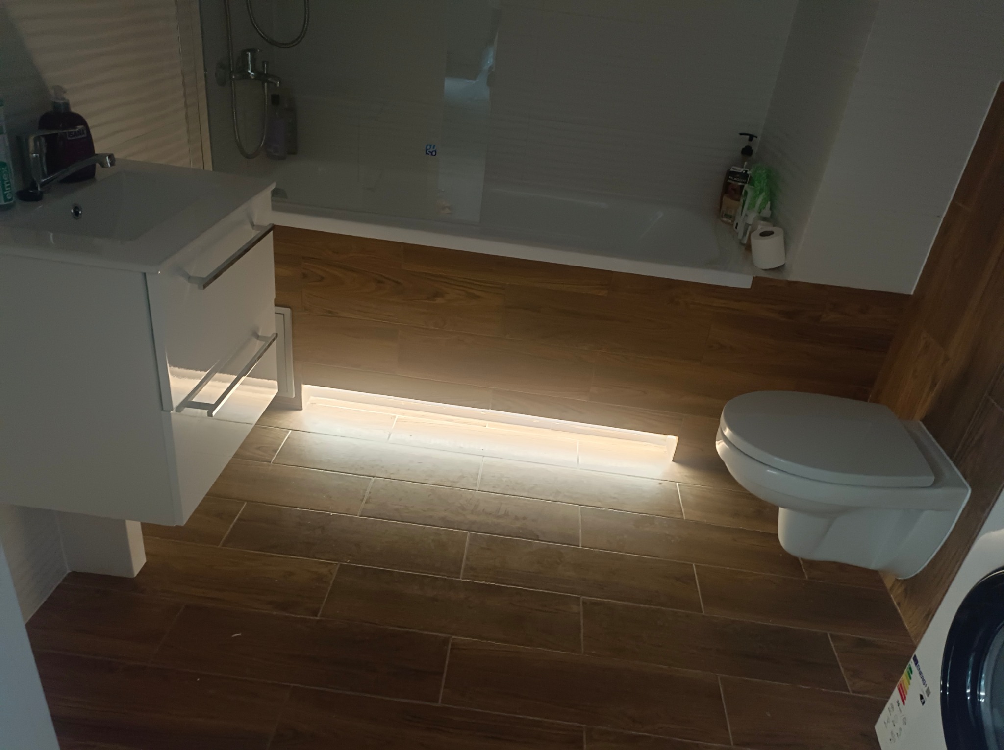 Nowoczesna łazienka z drewnopodobnymi płytkami, podświetleniem pod szafką umywalkową i wiszącą toaletą. Widoczna kabina prysznicowa i pralka.