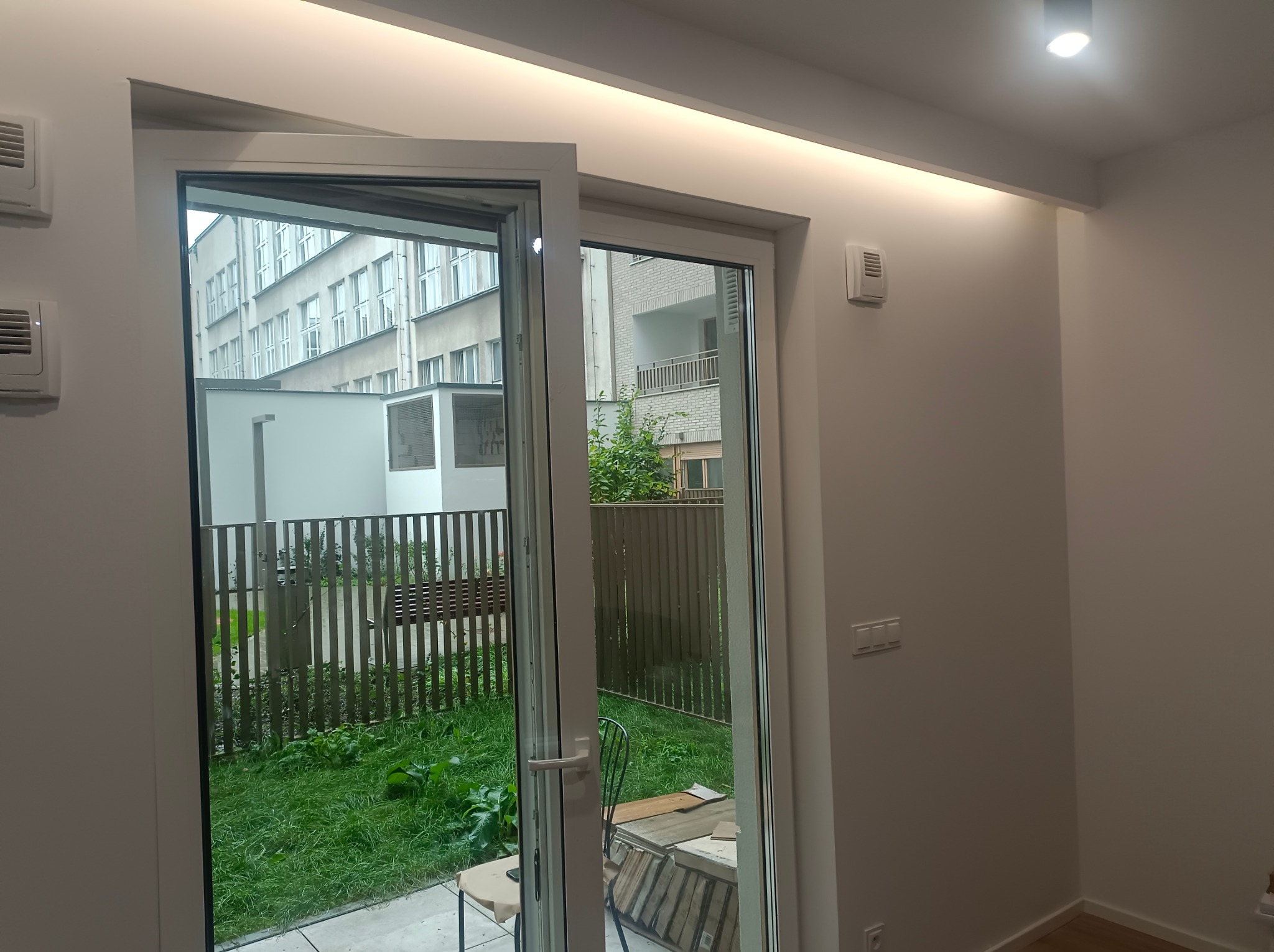 Otwarty balkon z widokiem na zielony ogród i budynek. Białe ramy okienne, oświetlenie sufitowe. Widoczne materiały budowlane.