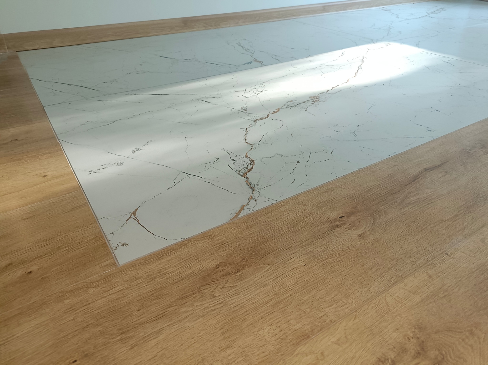 Połączenie podłogi: imitacja drewna i płytki imitujące marmur z delikatnym wzorem. Widoczne listwy przypodłogowe. Ujęcie z niskiego kąta.