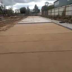 Przygotowany teren pod budowę z wyrównanym piaskiem i ułożonymi betonowymi płytami w tle widoczne drzewa i budynek przemysłowy.