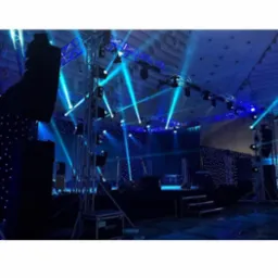Scena z metalową konstrukcją oświetleniową i reflektorami w kolorze niebieskim, ciemne tło z dekoracyjnymi światełkami, widoczne skrzynie transportowe na sprzęt.