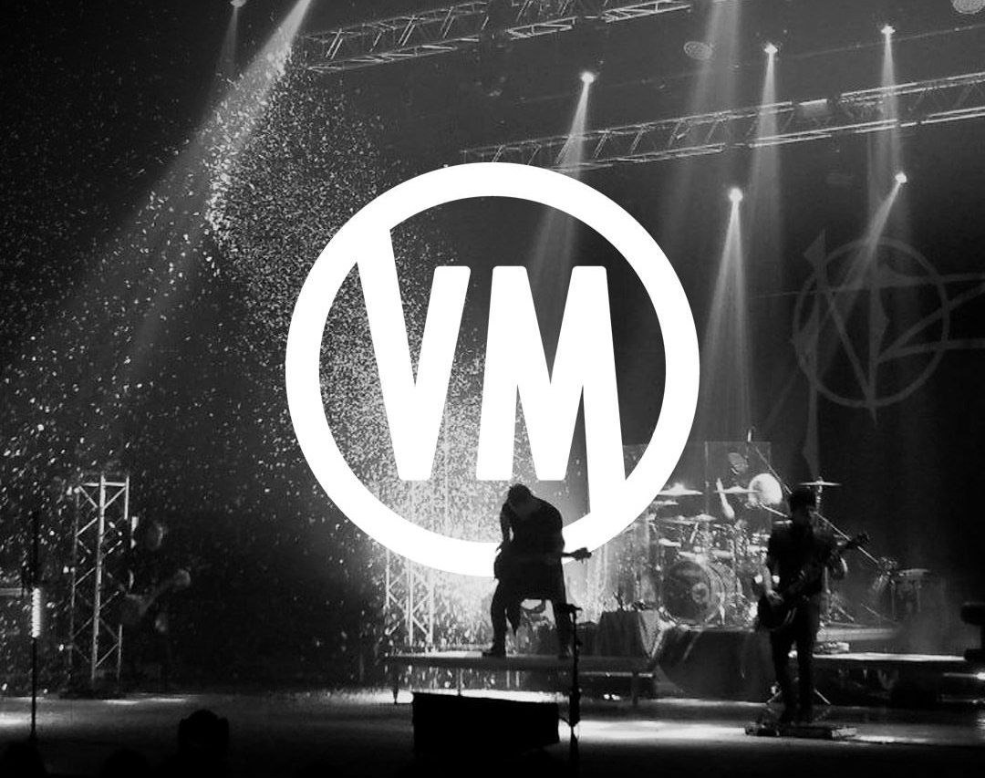 Czarno-białe zdjęcie zespołu muzycznego na scenie podczas występu, z reflektorami i efektem deszczu iskier, na pierwszym planie białe logo 'VM'.