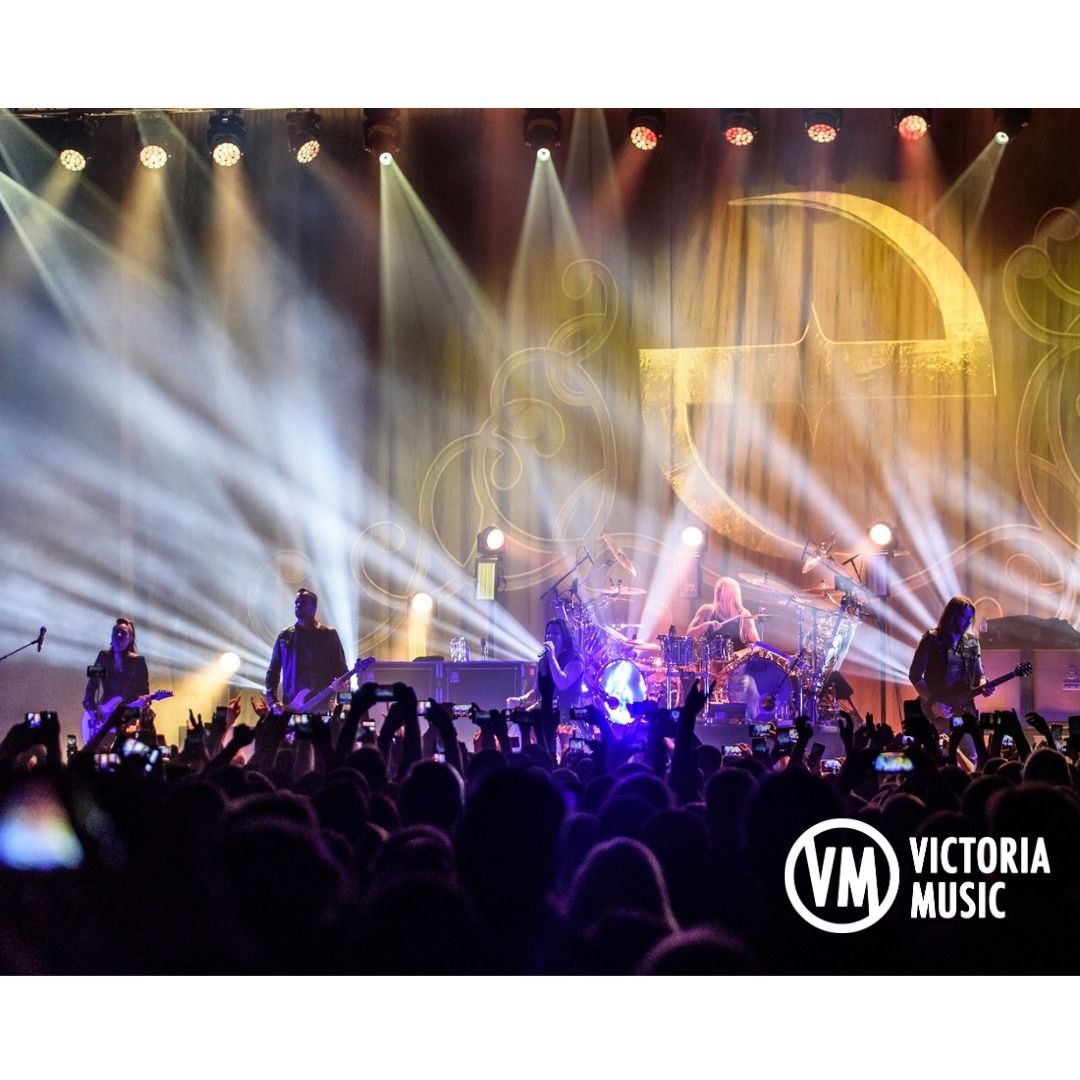 Zespół muzyczny na scenie podczas koncertu, oświetlenie punktowe, tłum fanów z telefonami w dłoniach, dekoracja sceniczna z dużym, złotym ornamentem.