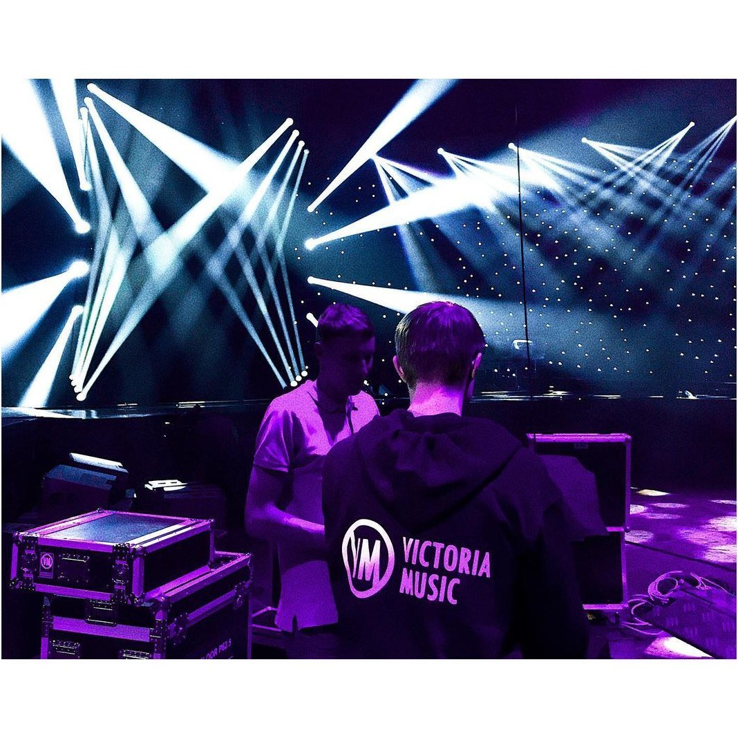 Technicy oświetleniowi przygotowują scenę z reflektorami i efektem gwiazd przed koncertem, widoczny sprzęt nagłośnieniowy i logo Victoria Music na bluzie jednego z techników.