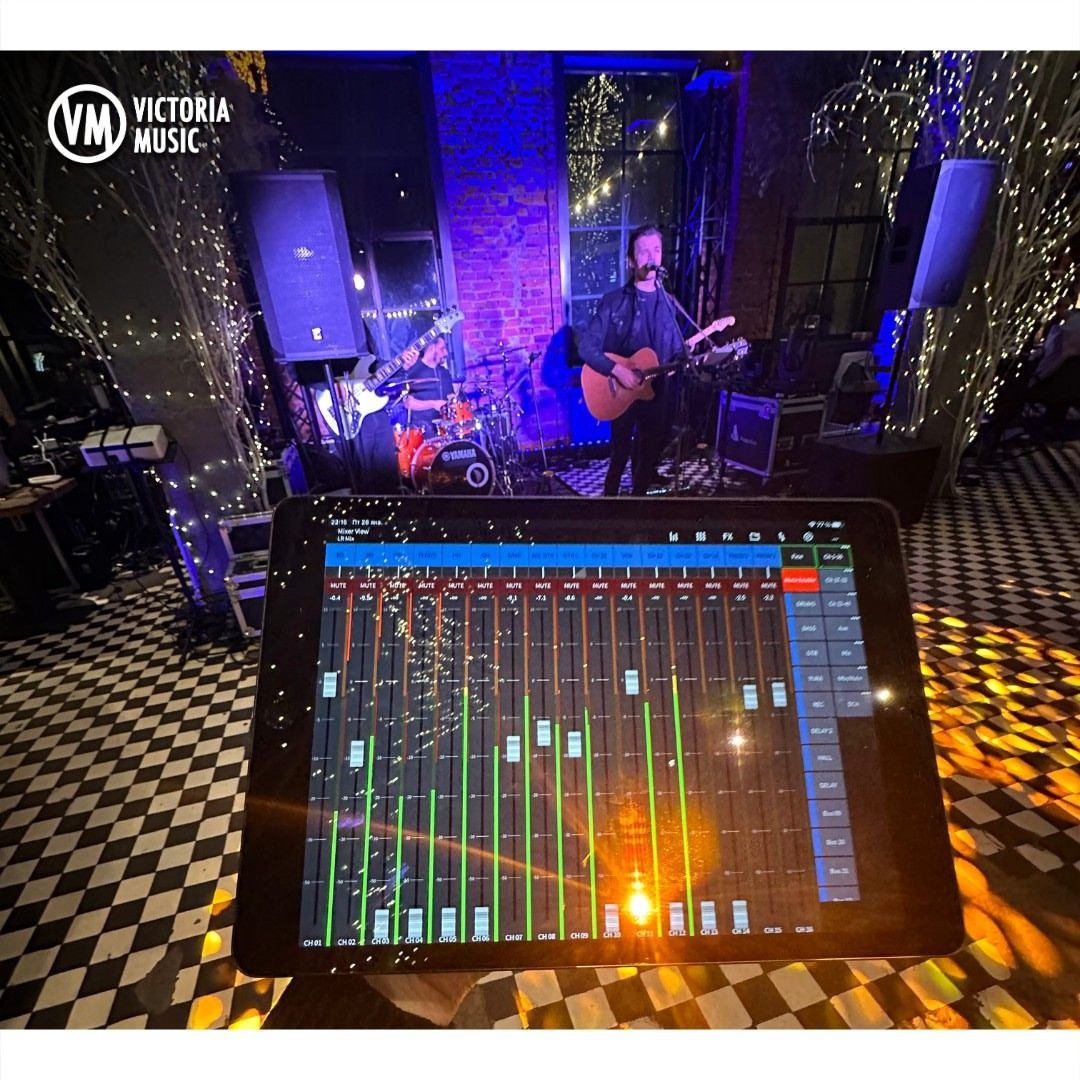 Zespół muzyczny grający koncert, widok zza konsoli mikserskiej na tablecie, oświetlenie sceniczne w odcieniach fioletu i bieli, logo zespołu w lewym górnym rogu.