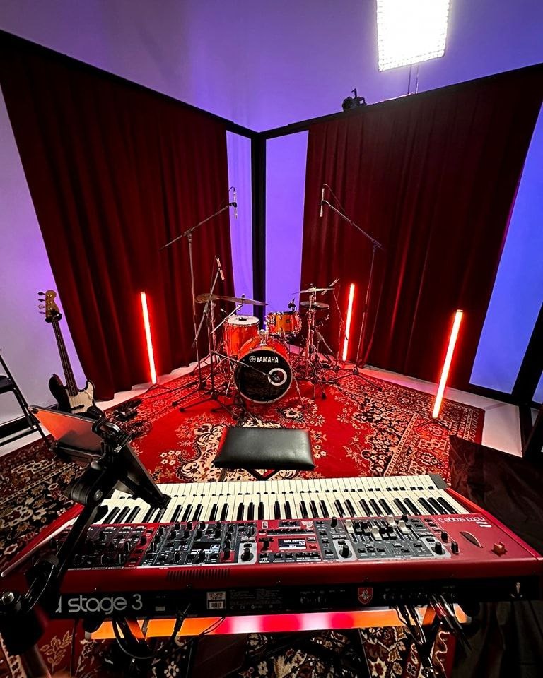 Ustawienie sceniczne z perkusją Yamaha, czerwonym dywanem, keyboardem Nord Stage 3 i pionowymi lampami LED, otoczone czerwonymi zasłonami i niebieskim oświetleniem.