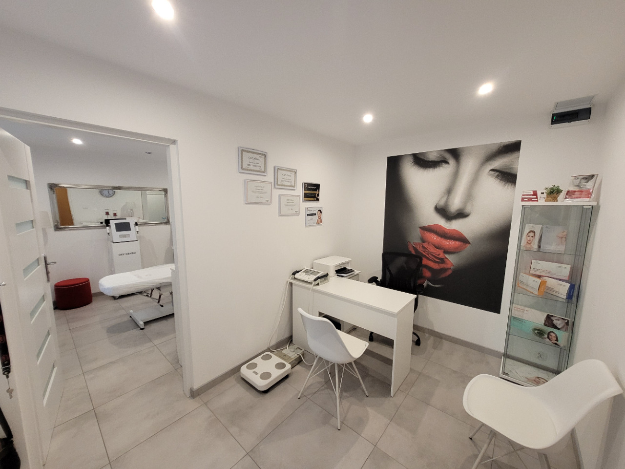 Jasne wnętrze gabinetu z biurkiem, wagą, plakatem kobiety z czerwoną szminką i regałem z kosmetykami, w tle widoczne łóżko zabiegowe i urządzenie.