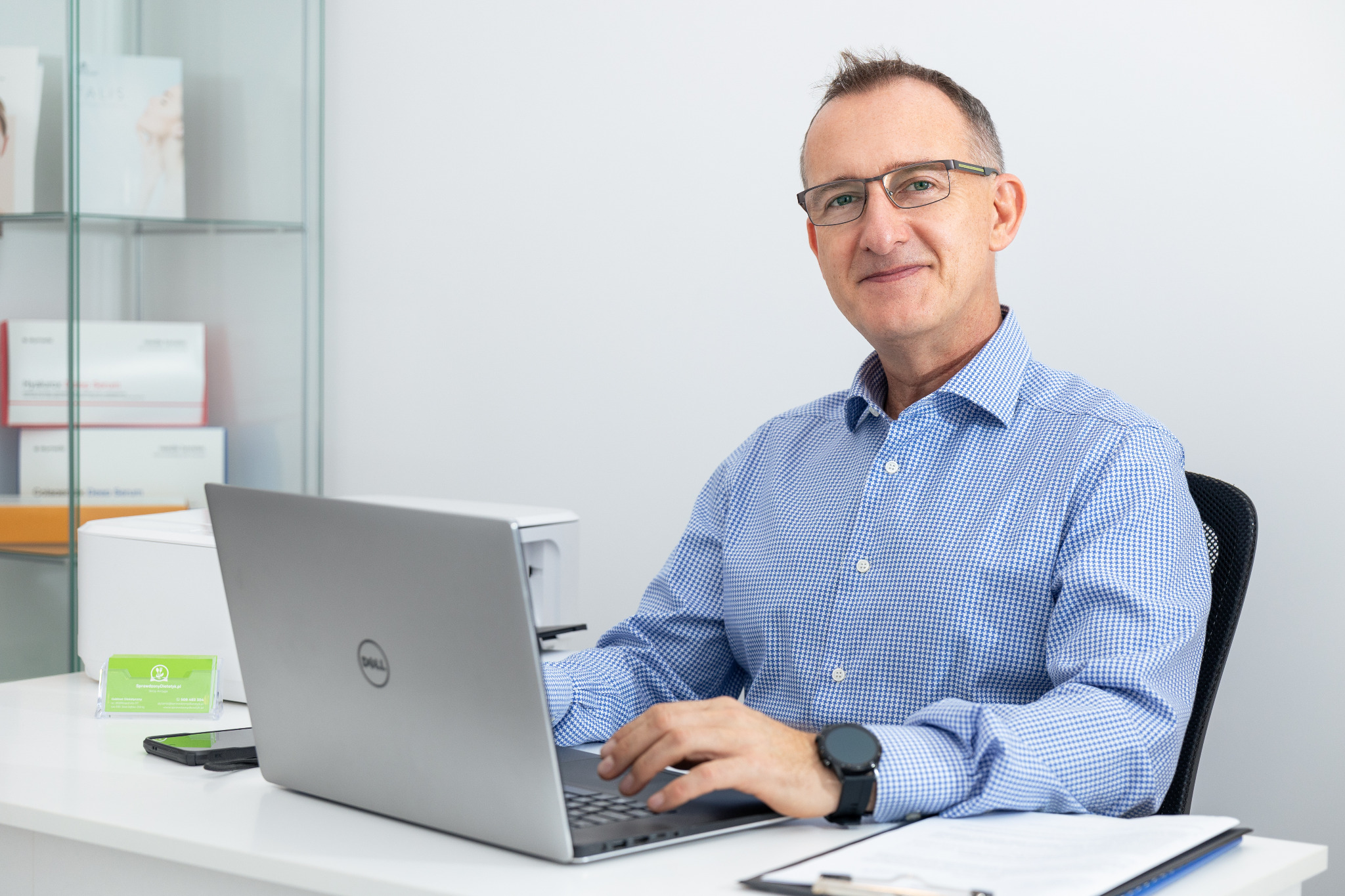 Mężczyzna w okularach siedzi przy biurku z laptopem Dell, obok wizytówka i dokumenty, w tle regał z kosmetykami.