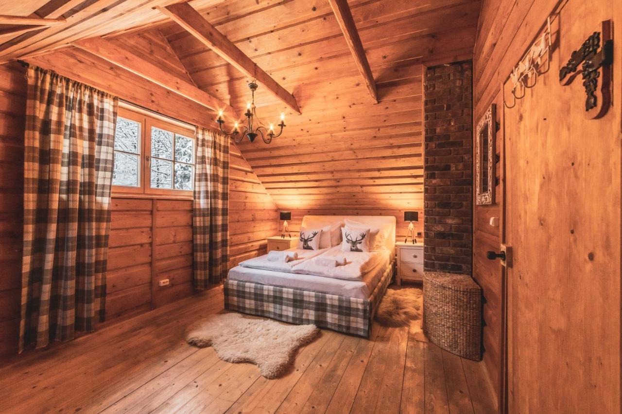 Drewniana sypialnia w stylu góralskim z łóżkiem w kratę, poduszkami z motywem jelenia i oknem z widokiem na ośnieżone drzewa.