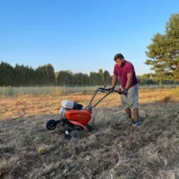 Mężczyzna w bordowej koszulce i szarych dresach obsługuje pomarańczowy glebogryzarkę Husqvarna na polu z suchą trawą, na tle drzew i błękitnego nieba.