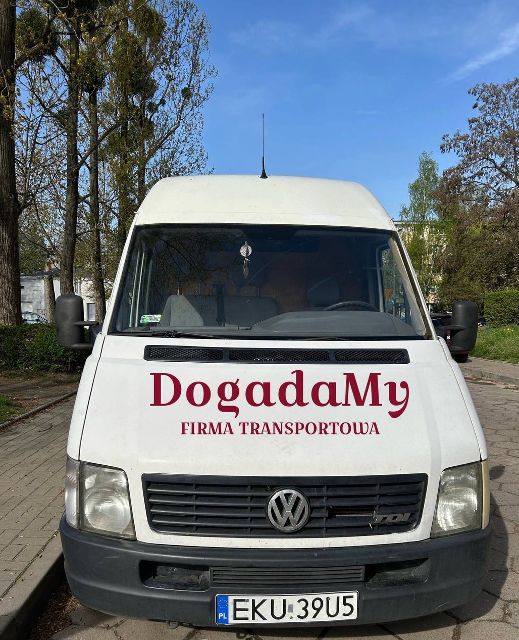 Biały samochód dostawczy Volkswagen z napisem 'DogadaMy FIRMA TRANSPORTOWA' na masce, zaparkowany na chodniku, z widocznymi drzewami w tle i niebieskim niebem.