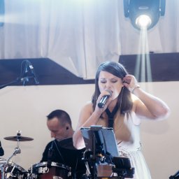 Joanna Herliczek - Wokalistka śpiewa do mikrofonu na scenie podczas występu, w tle perkusista i oświetlenie sceniczne.