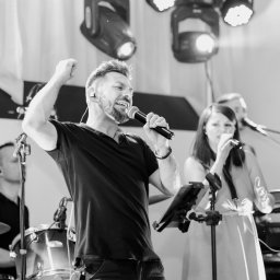 Joanna Herliczek - Czarno-białe zdjęcie zespołu muzycznego podczas występu na scenie, wokalista z mikrofonem i uniesioną ręką, w tle perkusja i drugi wokalista.