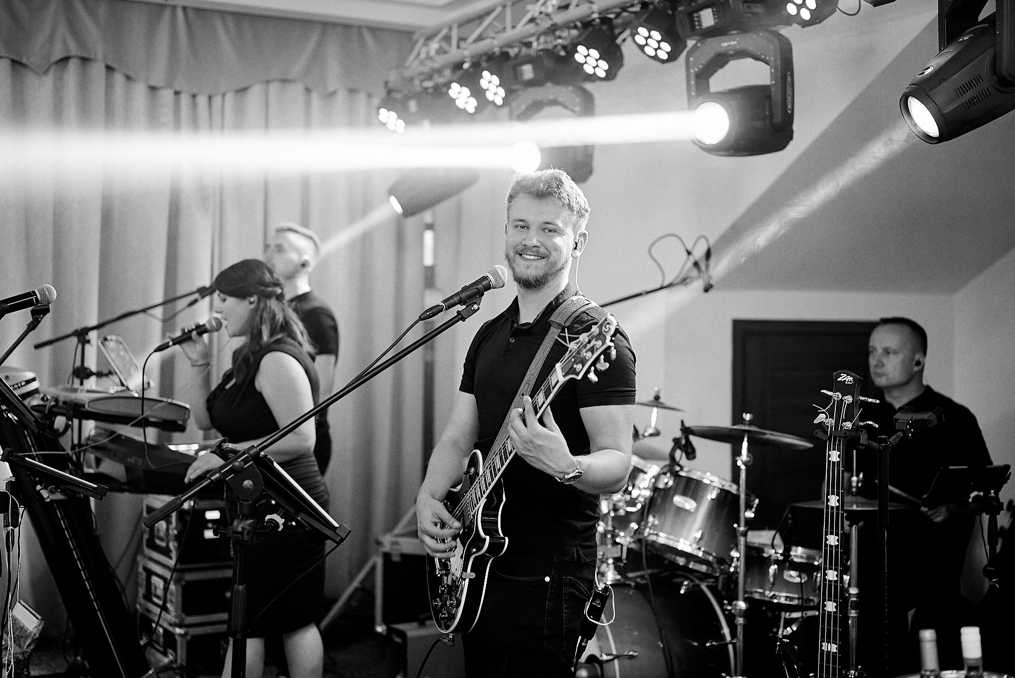 Czarno-białe zdjęcie uśmiechniętego gitarzysty z brodą, grającego na gitarze elektrycznej podczas występu na scenie z zespołem, oświetlonej reflektorami