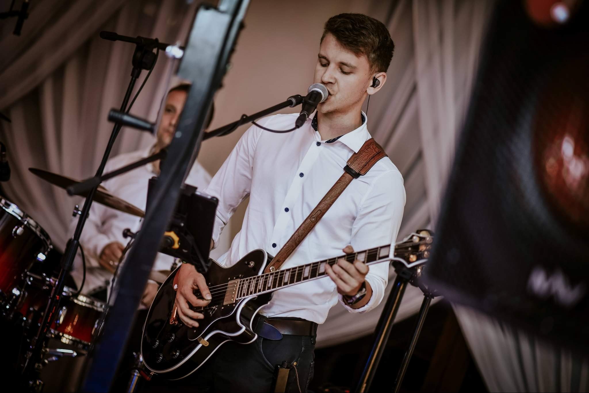 Wokalista z gitarą elektryczną śpiewa do mikrofonu na scenie, w tle perkusista i białe draperie.