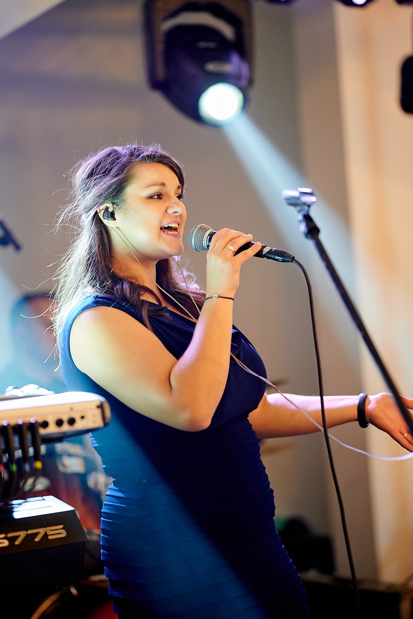 Wokalistka w niebieskiej sukience śpiewa do mikrofonu na scenie, obok widoczny keyboard Yamaha S775, oświetlona reflektorem.