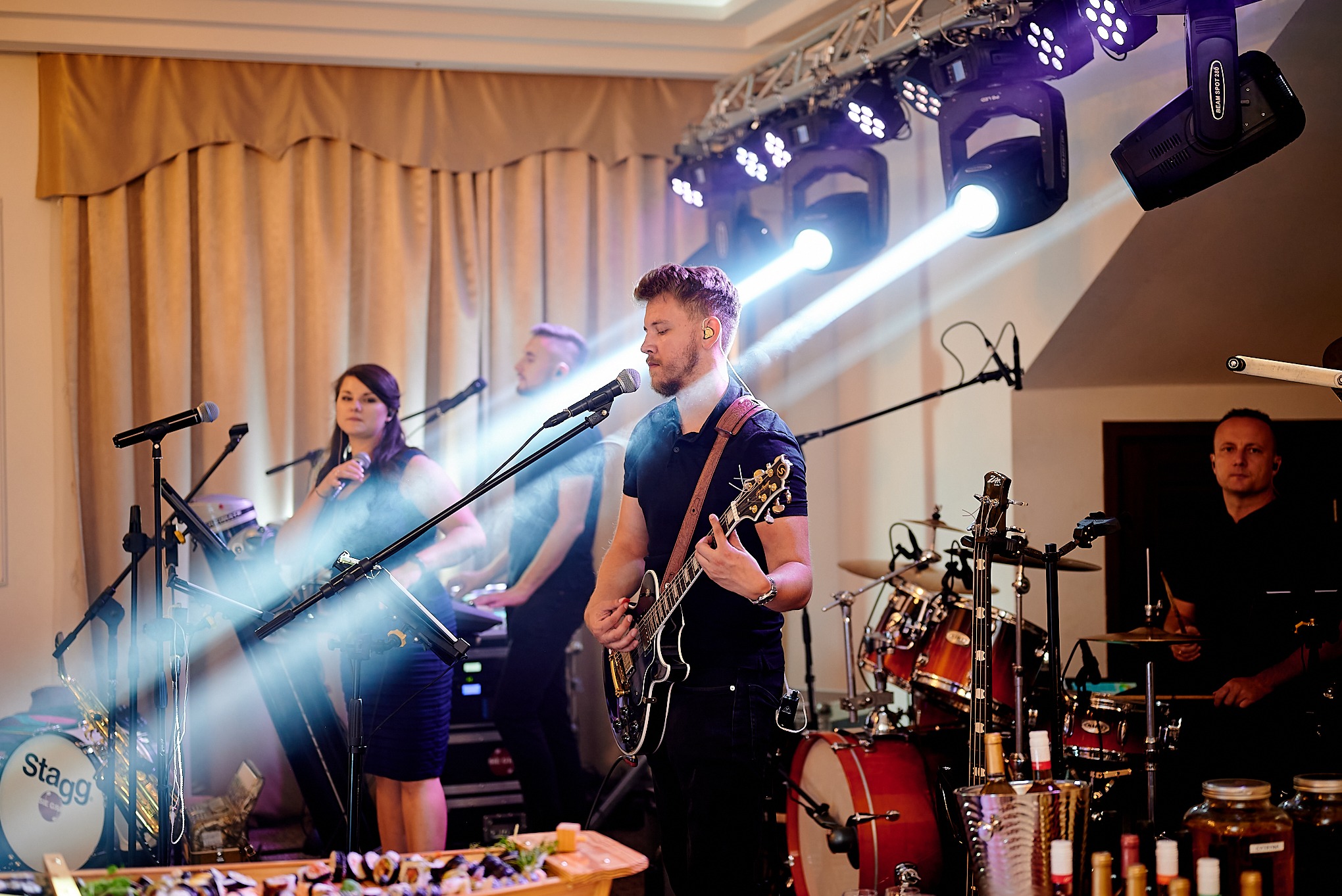 Zespół muzyczny grający na żywo podczas wydarzenia, oświetlony reflektorami, z widocznym cateringiem sushi na pierwszym planie.