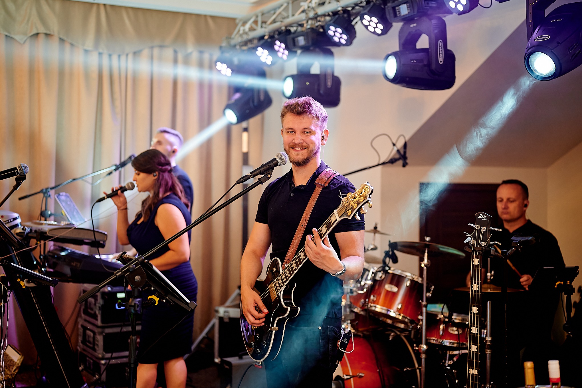 Uśmiechnięty gitarzysta z brodą w centrum kadru, w tle wokalistka, klawiszowiec i perkusista podczas występu na scenie z profesjonalnym oświetleniem.