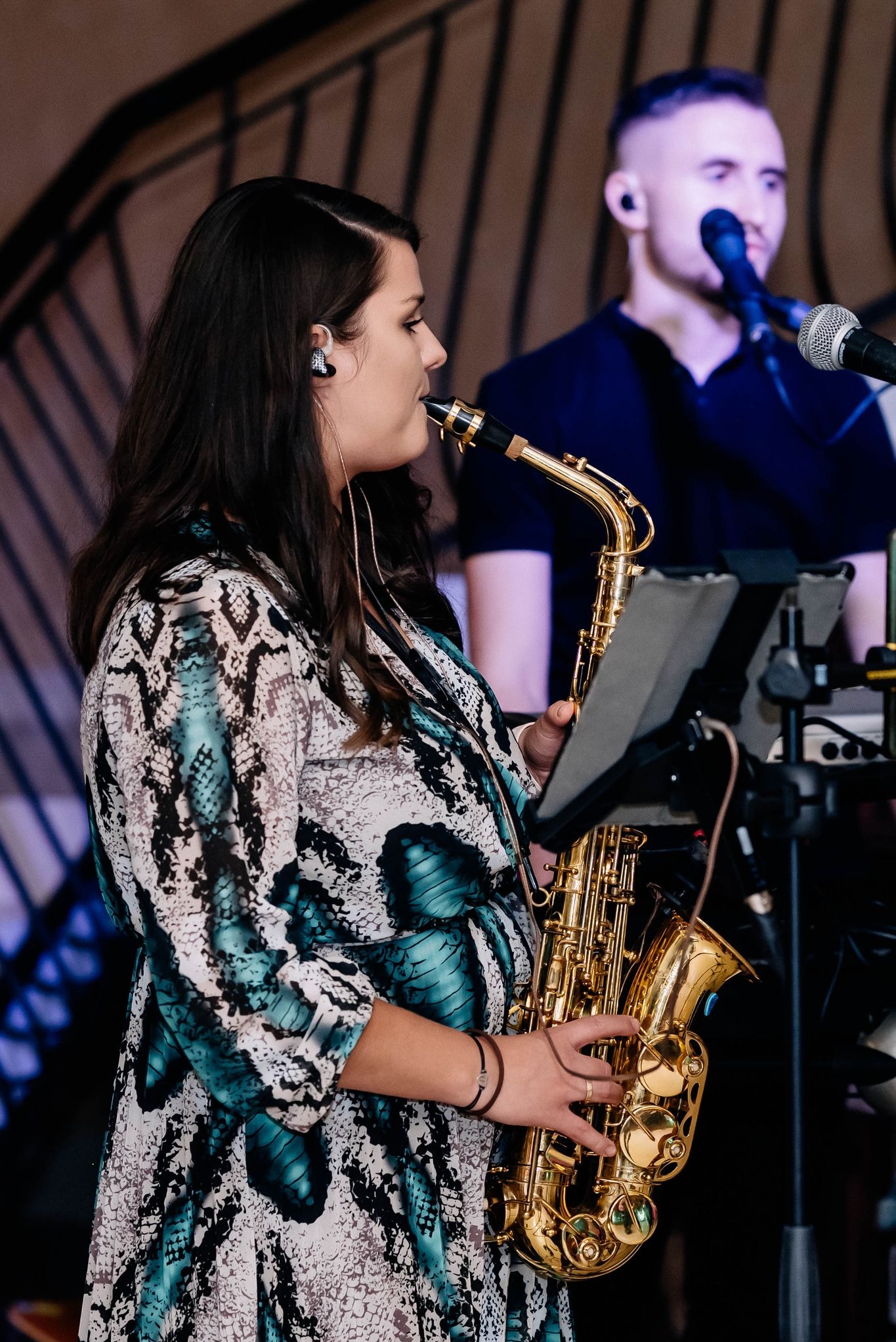 Kobieta w sukience w motywy zwierzęce gra na saksofonie, na scenie widoczny również wokalista z mikrofonem.