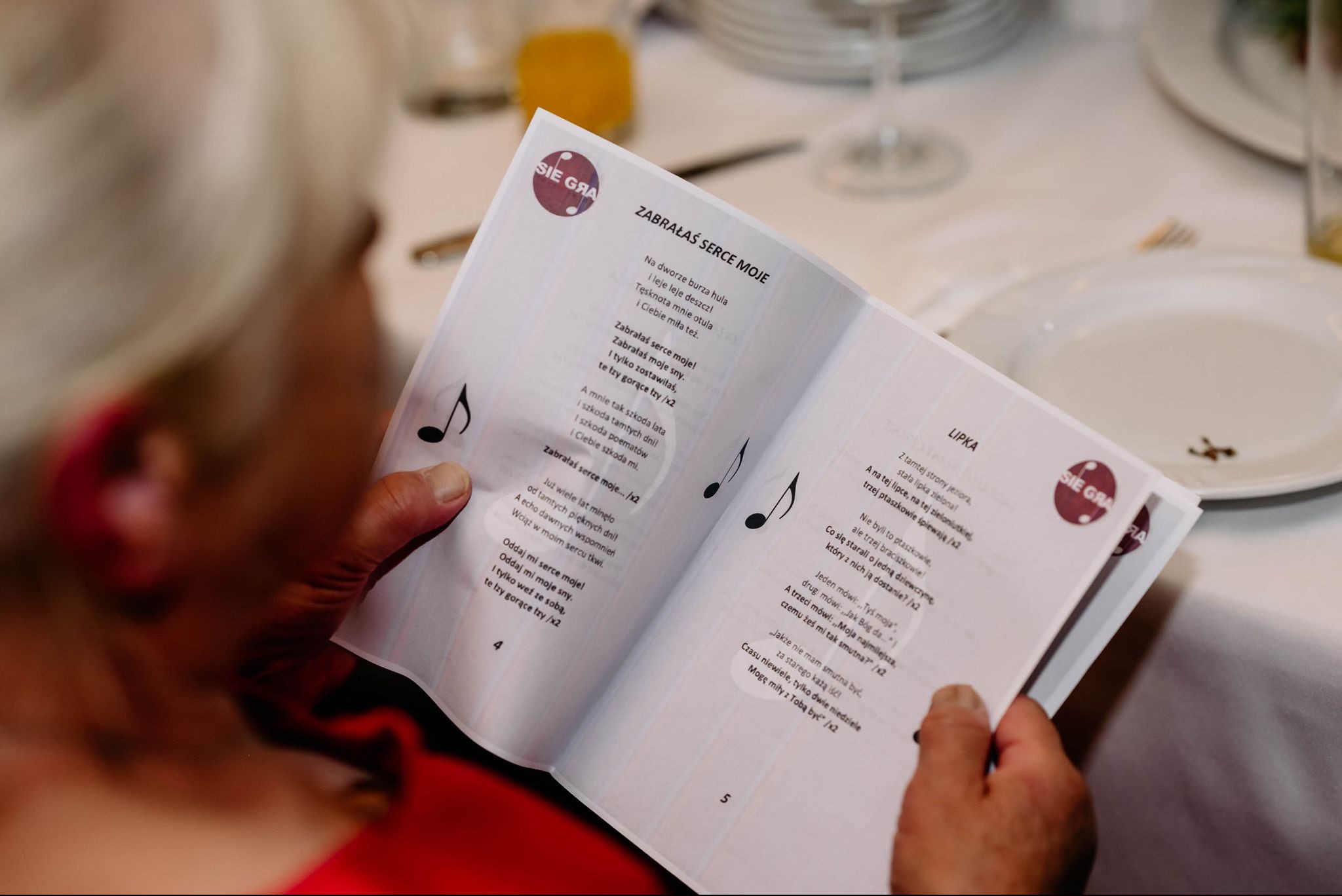 Osoba trzyma w rękach kartkę z tekstem piosenki 'Zabrałaś serce moje' i 'Lipka', widoczne nuty i logo 'Sie Gra', sugerujące śpiewnik lub repertuar na imprezę.